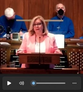 Linda Lotz standing at a lectern at Towson Presbyterian, with two members of the church choir in the background